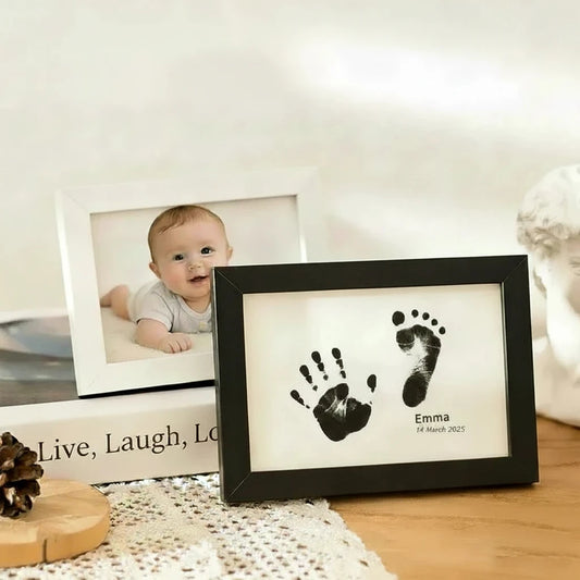 Two light wood frames on a tabletop, one showing a baby photo and the other displaying black baby footprint prints on a white insert with the name Emma. Text at the top reads 'Baby Footprint Photo' and 'Frame Record Beautiful Moments in Life.