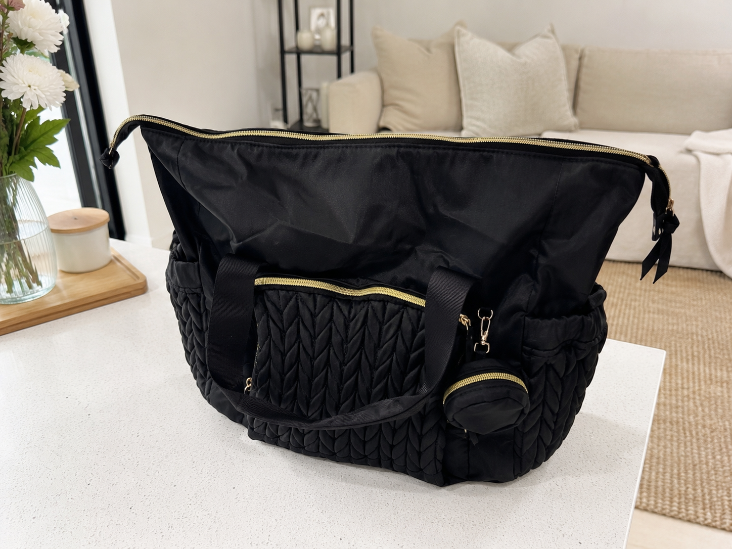 A black quilted duffel bag with gold zipper accents sits on a white table in a bright, modern living room setting.