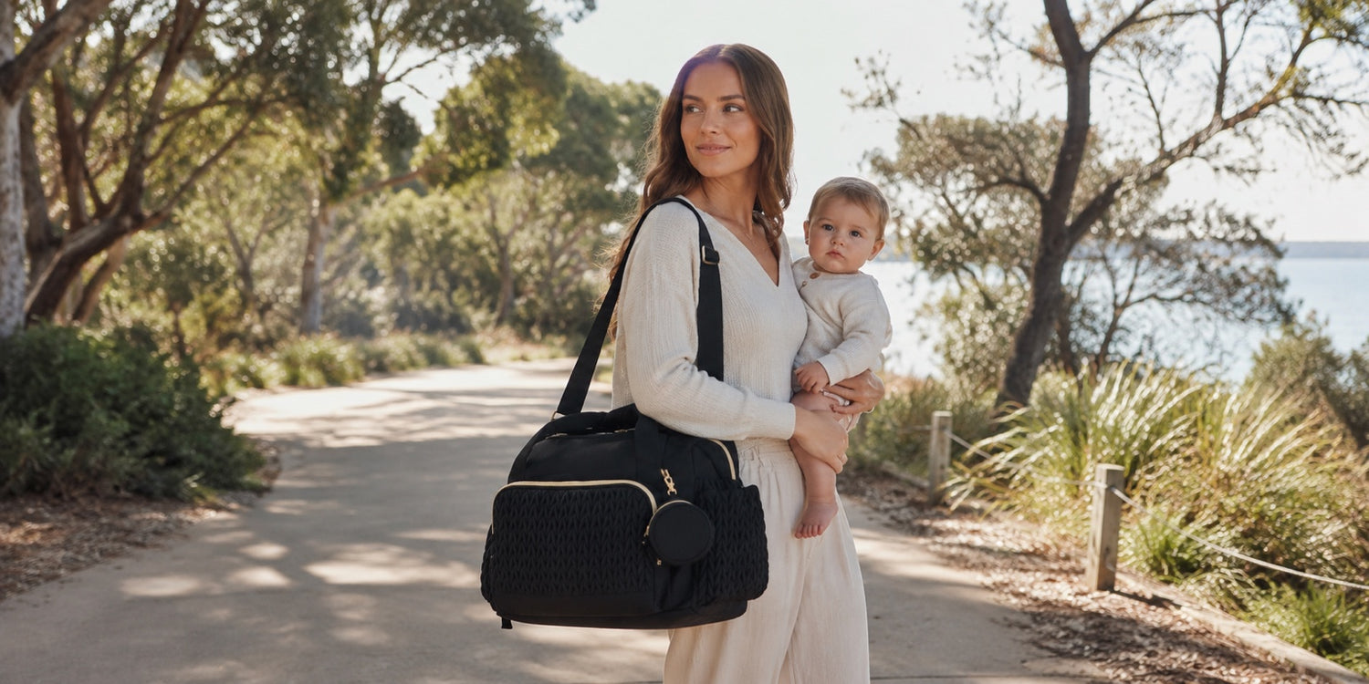 Woman holding a baby and a black bag on a path with trees and grass in the background